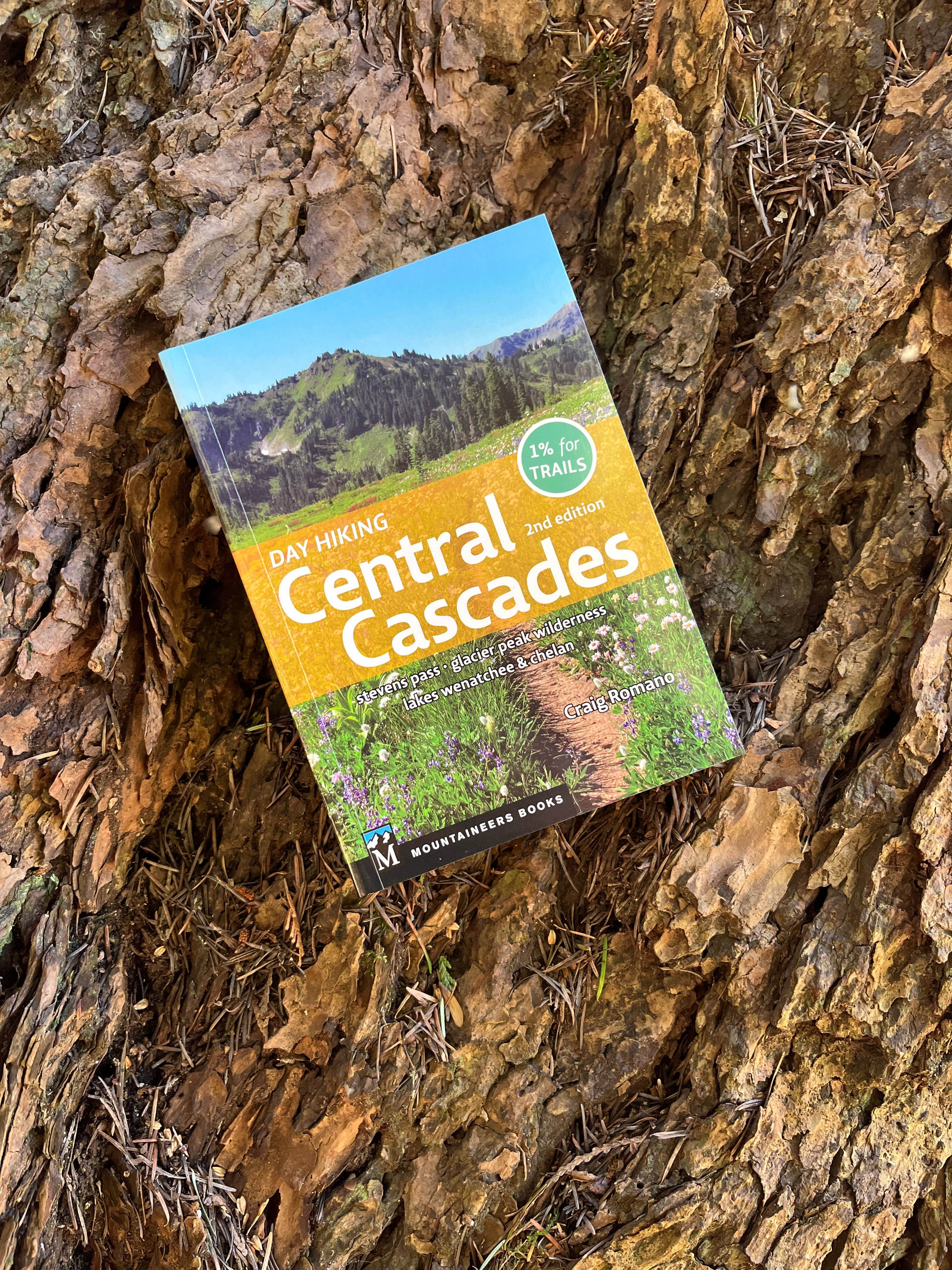 Front cover of "Day Hiking Central Cascades 2nd Edition" by Craig Romano background picture of a meadow and mountain trail in Cascades. 