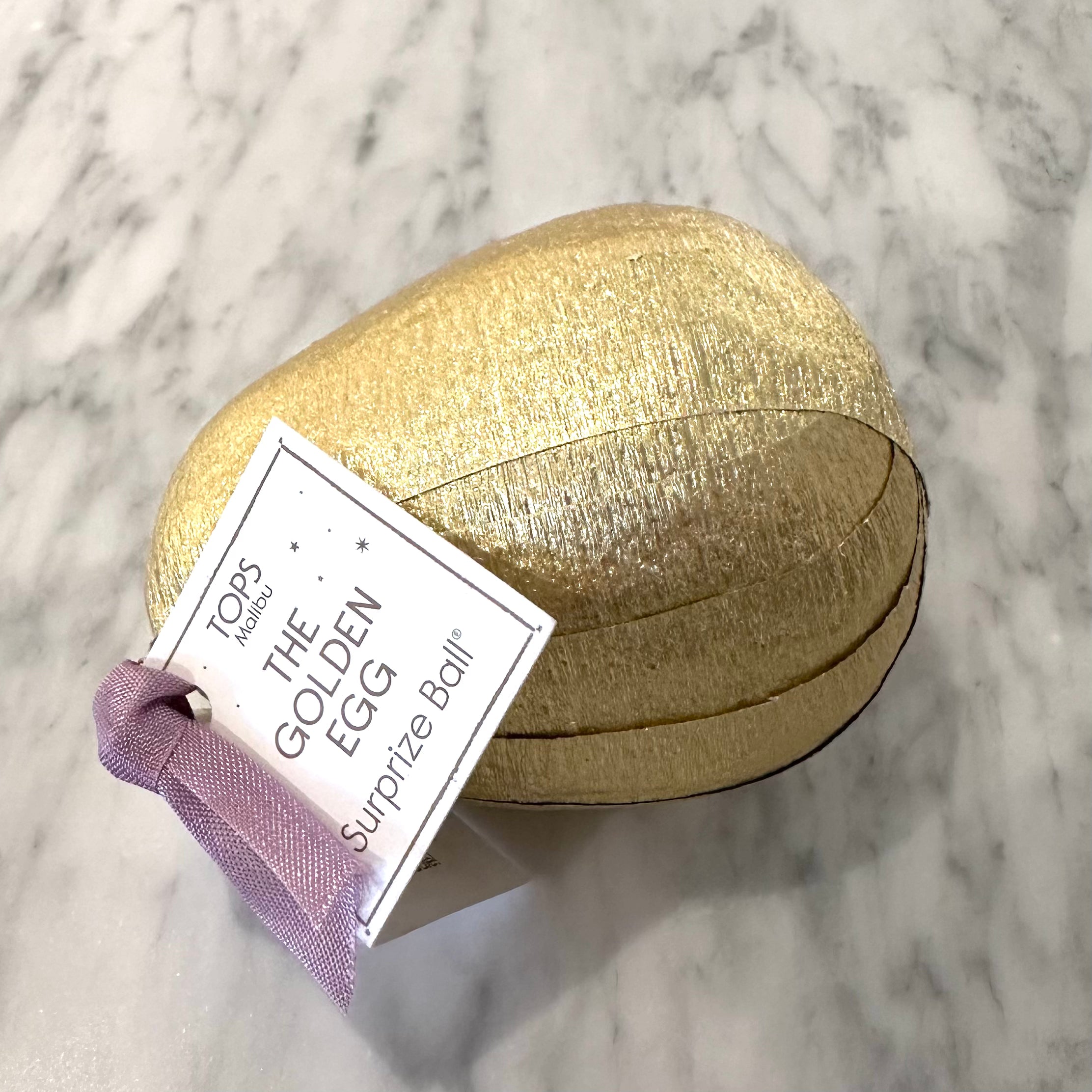 Golden surprise ball in egg shape, white tag with black writing states "The Golden Egg Surprize Ball"