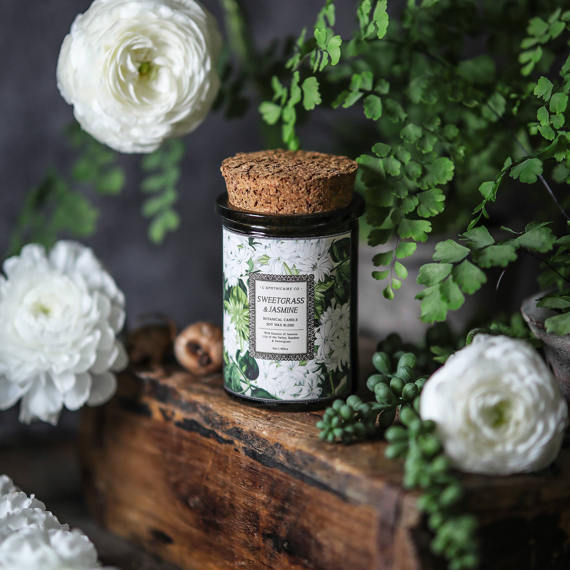 Candle inside green glass jar with cork lid, white jasmine flowers with green leaves on label.  Label states "sweetgrass and jasmine" as the scent. 